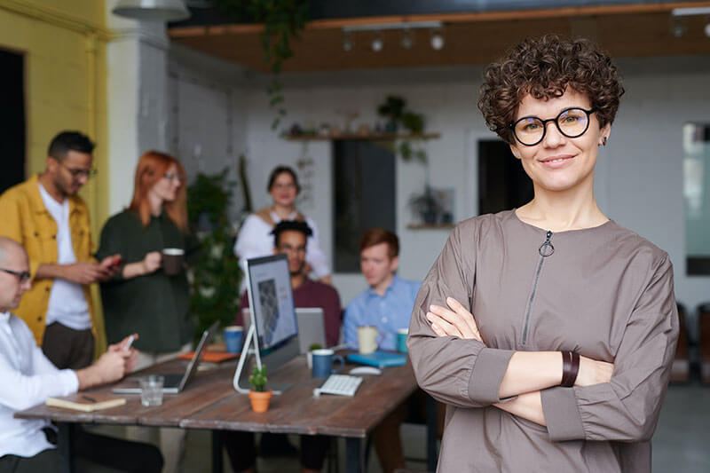 woman with curly hair stands confidently with her arms crossed in front of people around a desk